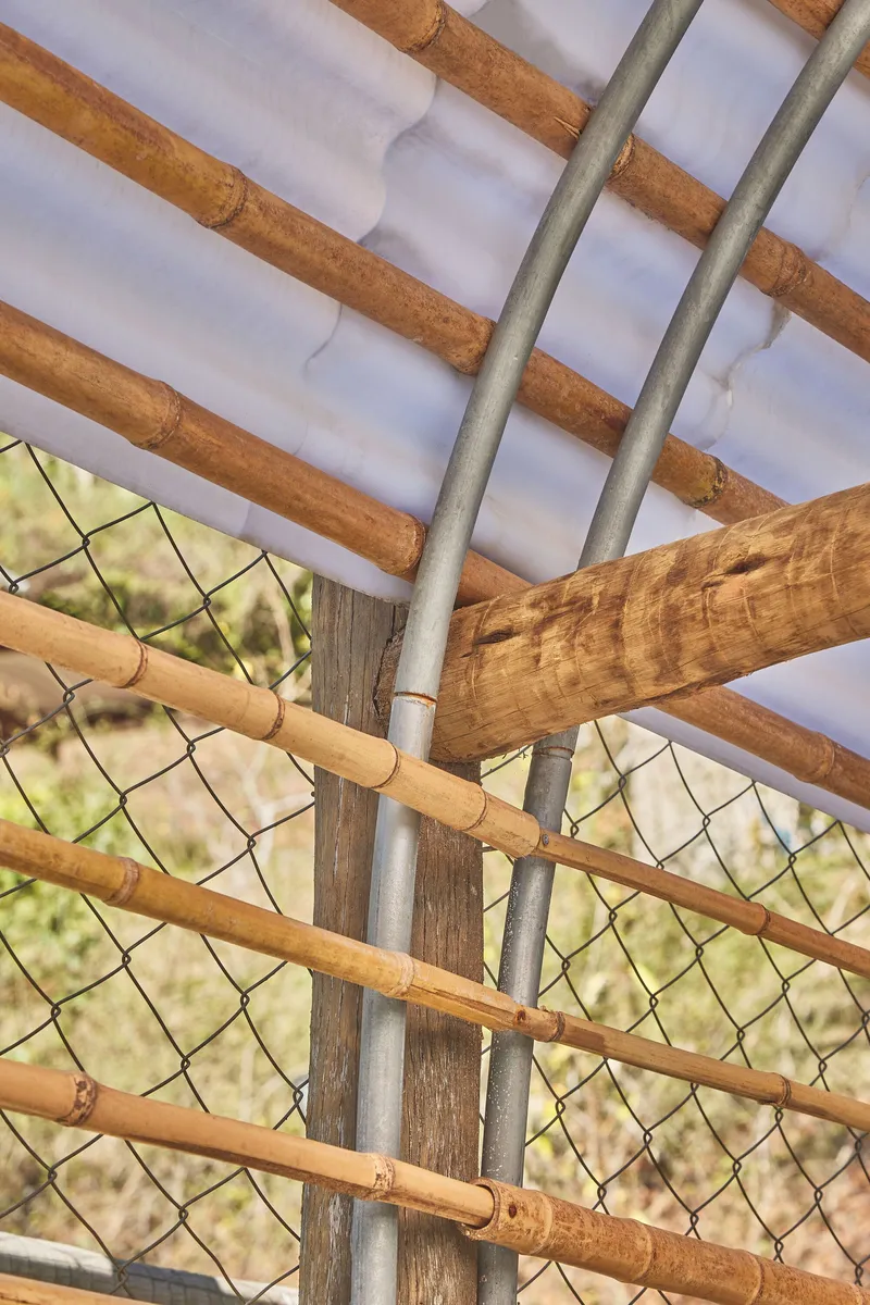 Detail of bamboo poles, chain-link mesh, and galvanized pipe junction at the pavilion corner