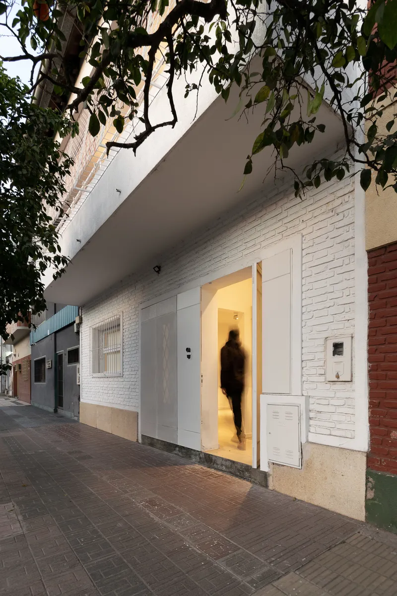 White painted brick facade with open door revealing illuminated interior at dusk with a blurred figure