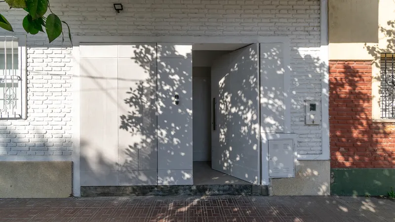 Street facade with pivoting white steel doors partially open, casting dappled tree shadows across the brick wall