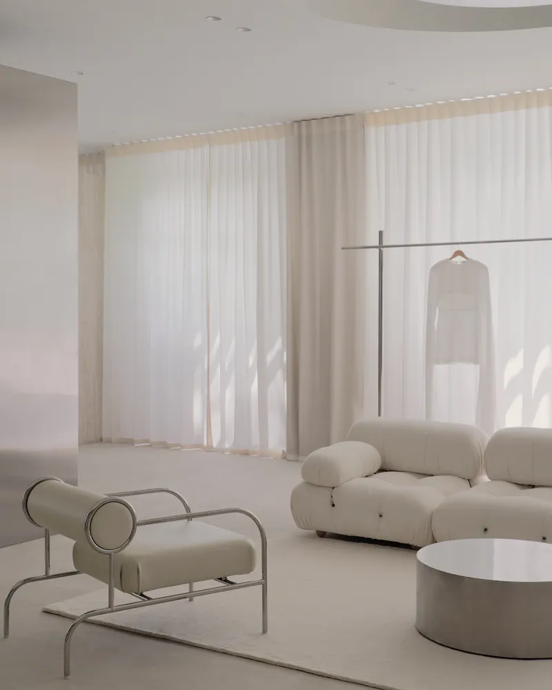 Tubular steel lounge chair and modular sofa beneath layered sheer and solid curtains in daylight
