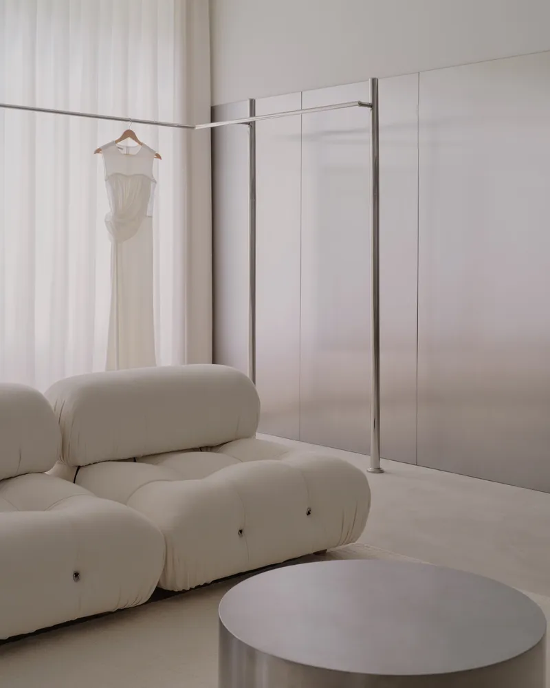 Dressing area with freestanding steel clothing rack, hanging white dress, and cushioned seating below sheer curtains