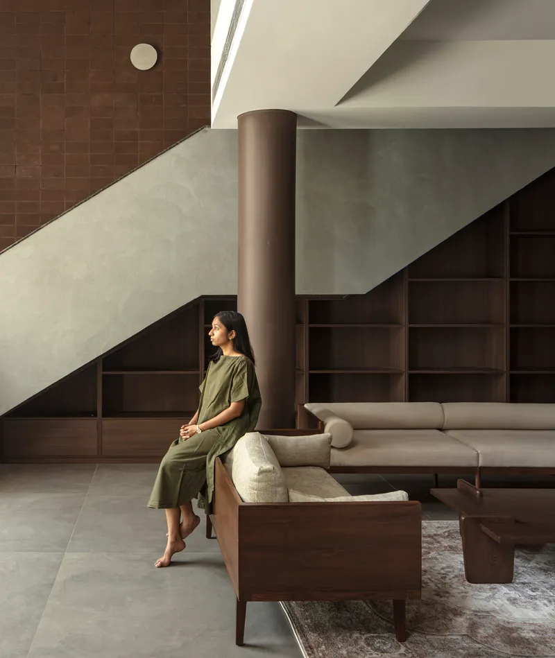 Figure seated beside the staircase column with shelving and sofa in the background