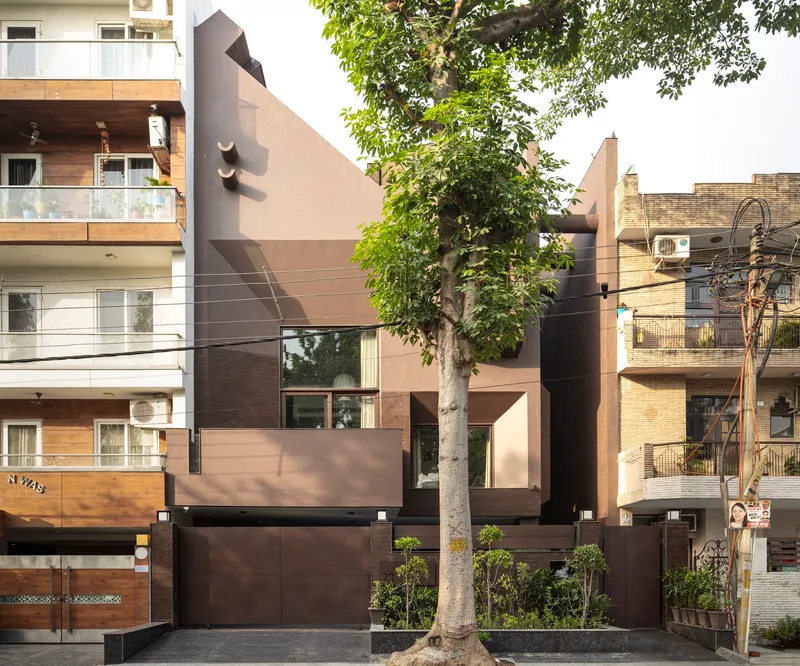 Street facade with layered brown and beige panels framed by a mature tree and neighboring buildings