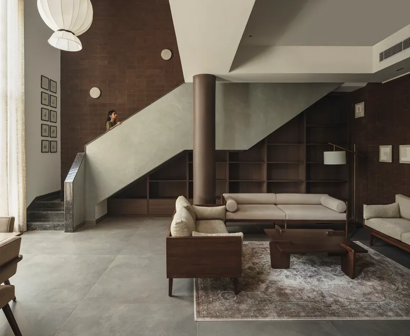 Double-height living space with pale staircase, cylindrical column, timber seating, and pendant light overhead