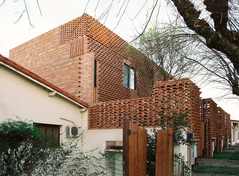 Street view of the perforated brick facade rising above adjacent houses in bare winter branches
