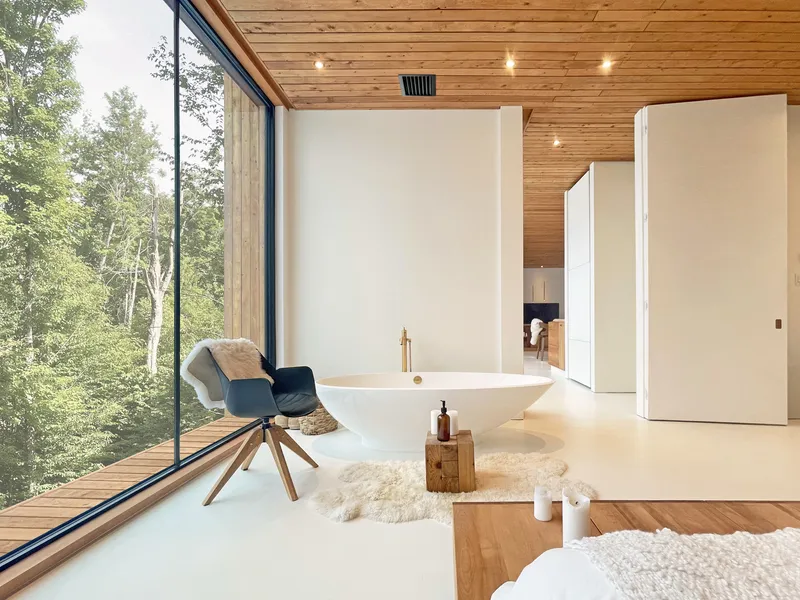 Freestanding bathtub on white flooring beneath timber ceiling with full-height glazing to forest beyond
