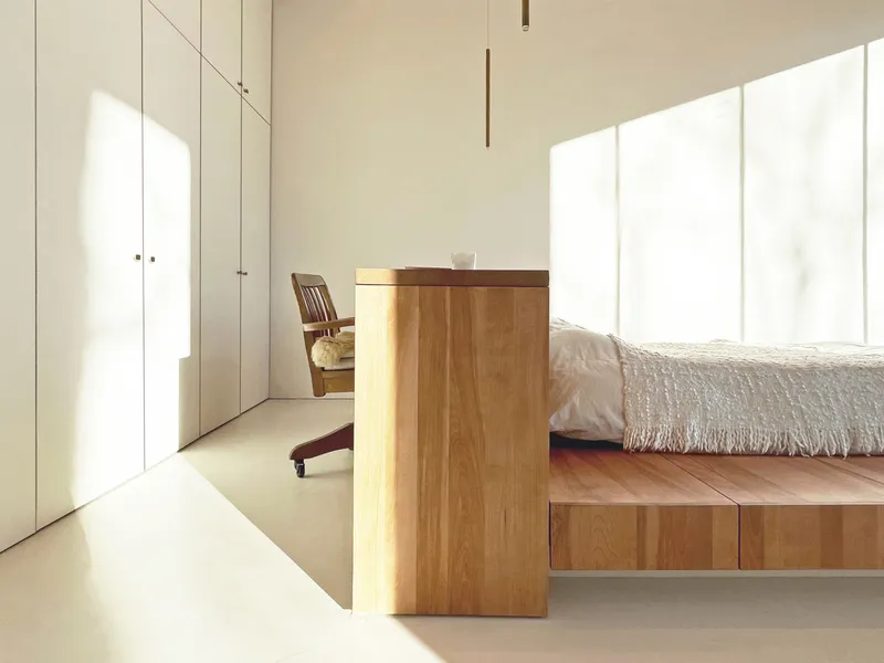 Minimalist bedroom with timber platform bed and floor-to-ceiling white storage cabinetry bathed in natural light