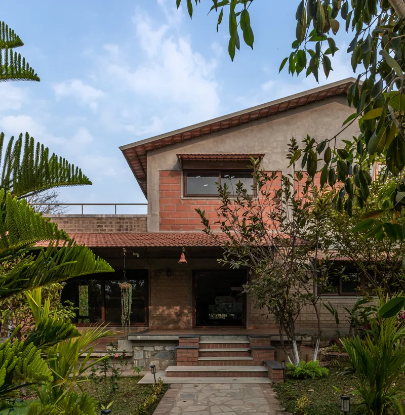 Front facade with red brick and stone cladding framed by palm fronds and ferns beneath a cloudy sky