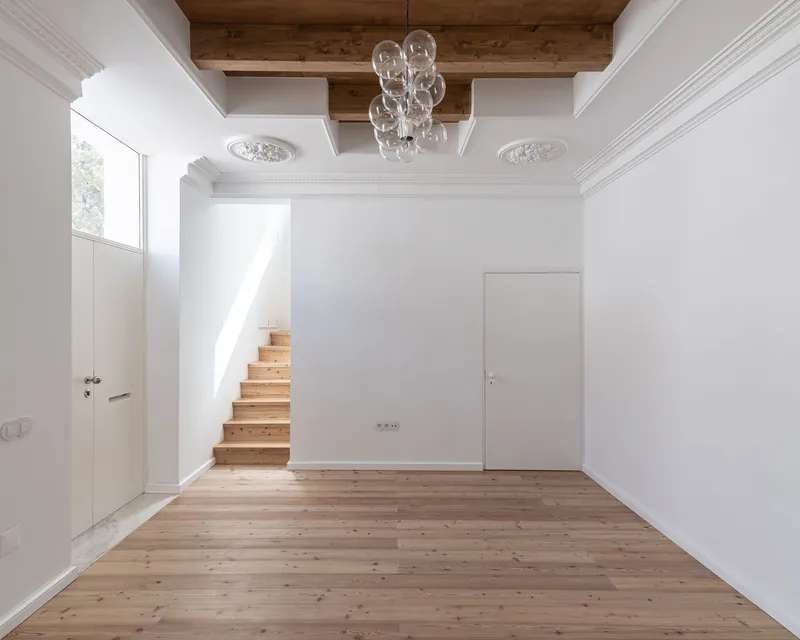 White interior with timber staircase emerging from the wall below exposed timber ceiling beams