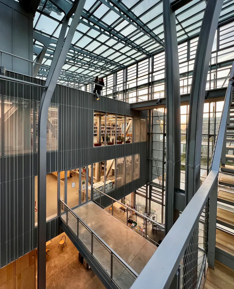 Multi-story atrium with corrugated metal volumes suspended between exposed steel structural framework and glass ceiling