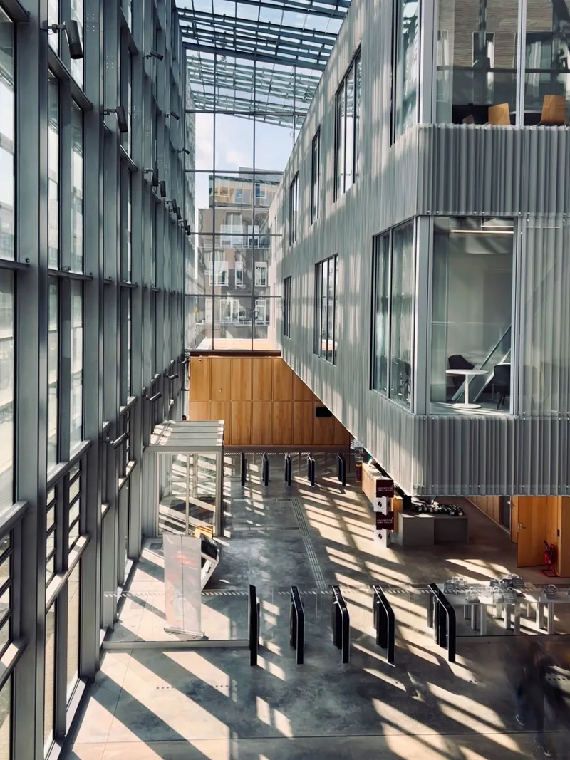 Glazed atrium corridor with steel and glass roof casting shadows across the concrete floor at midday