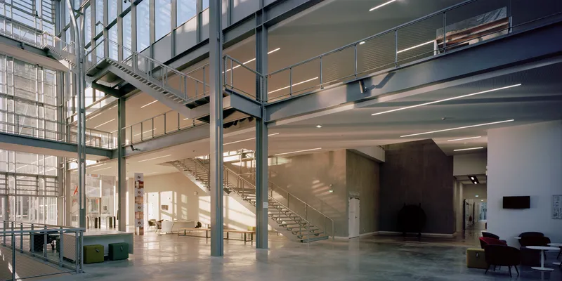Double-height lobby with exposed steel columns, glass partitions and a floating staircase under linear ceiling lights