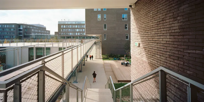 Elevated pedestrian ramps with mesh safety barriers winding between brick-clad volumes and courtyard planting