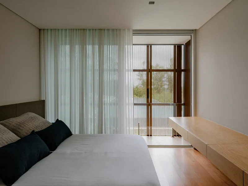 Bedroom with vertical timber screen filtering daylight beside sheer curtains and built-in wood desk