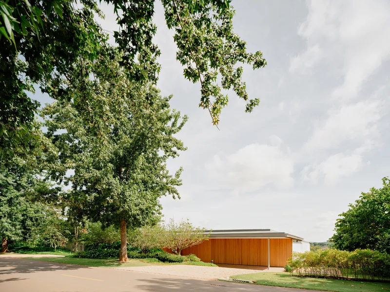 Horizontal timber facade and flat roof cantilevering over a gravel drive edged by lawn and trees