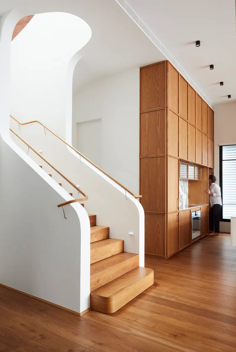 Timber staircase with brass handrail ascending beside full-height oak cabinetry wall with integrated appliances