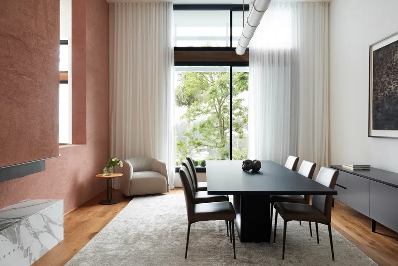 Dining room with sheer curtains filtering light from garden-facing windows and rammed earth fireplace wall