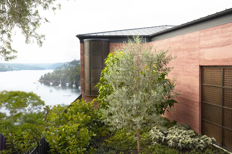 Rammed earth facade with planted slope and harbor view beyond the tree canopy