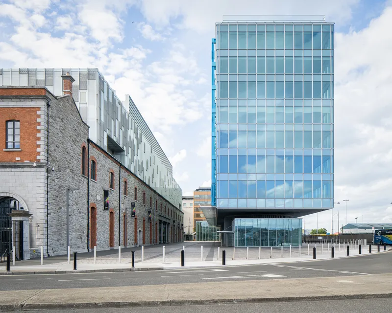 Street view of a cantilevered glass tower alongside a restored stone warehouse under clouds