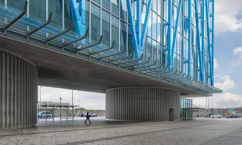 Underside of the cantilevered volume with vertical concrete fins and a pedestrian crossing the plaza