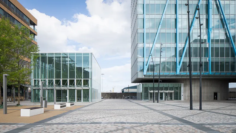 Paved plaza between glass pavilion and tower with blue structural bracing under scattered clouds