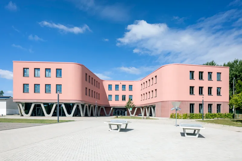Courtyard view of the pink rendered facade with white V-shaped structural columns and picnic tables