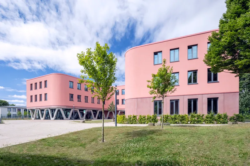 Pink facade with rounded corners and young trees in a lawn under scattered clouds