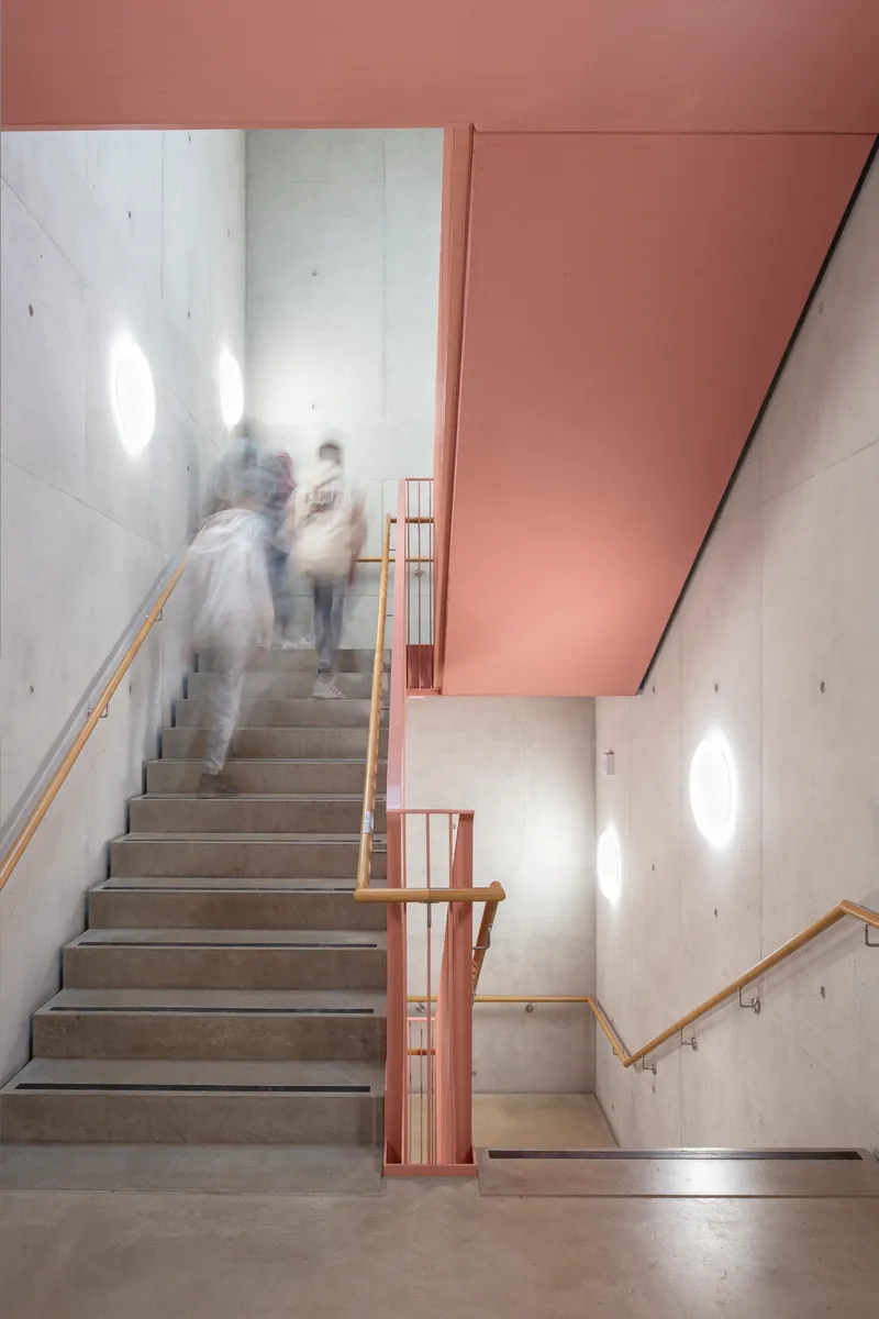 Concrete stairwell with wooden handrails and blurred figures ascending beneath a pink soffit