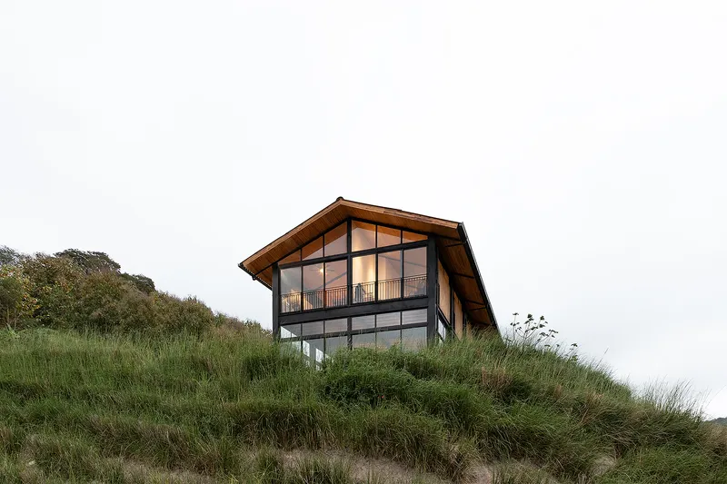 Two-story gabled facade with steel-framed glazing and timber cladding atop a grassy dune