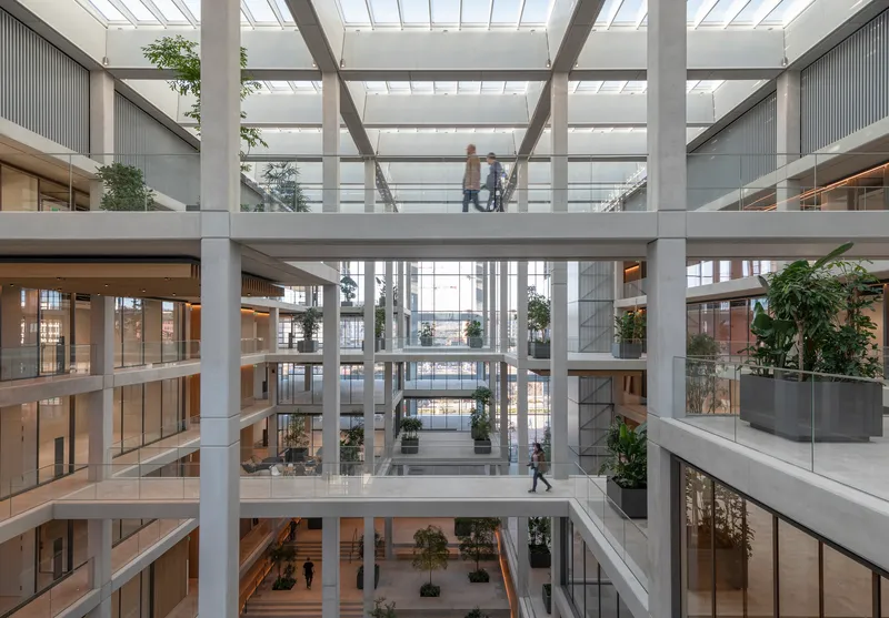 Multi-storey atrium with concrete beams, glass skylight and people crossing a central bridge