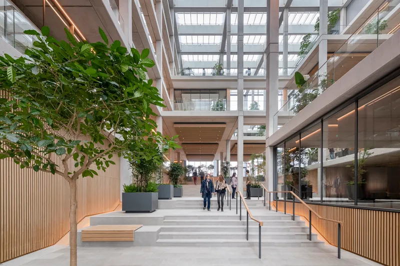 Interior stairway with timber benches and planted trees beneath a glass-roofed atrium with people ascending