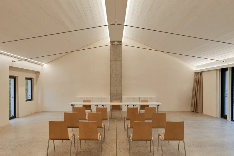 Meeting room with vaulted timber ceiling, central concrete column and rows of plywood chairs on terrazzo floor