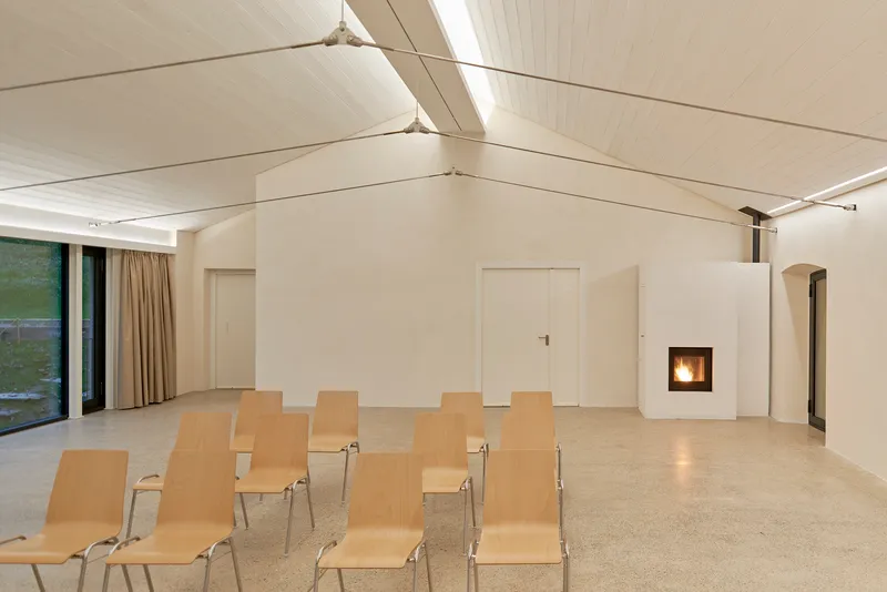Assembly space with gabled fabric ceiling, terrazzo floor, plywood seating, and freestanding fireplace with active flame