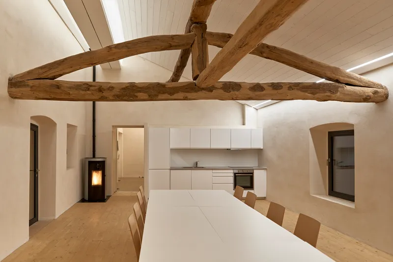 Dining area with exposed timber roof truss above white table and kitchen cabinetry along rear wall