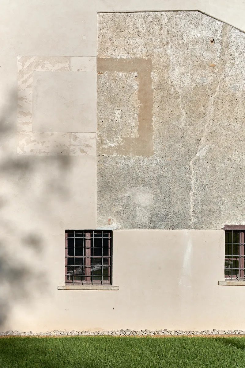 Layered plaster facade with patched repairs and two barred windows above a grass lawn