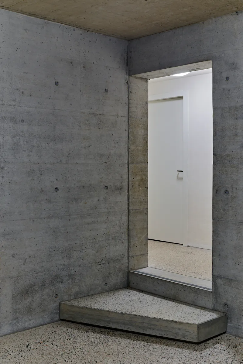Board-formed concrete entryway with stepped platform and white door opening in dim interior light