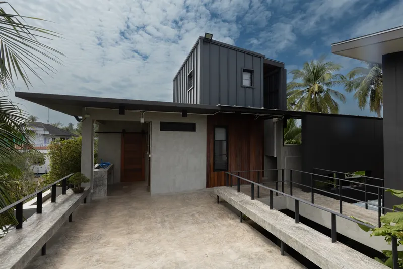 Upper-level terrace with concrete flooring and steel railings beneath a corrugated metal volume and palm canopy