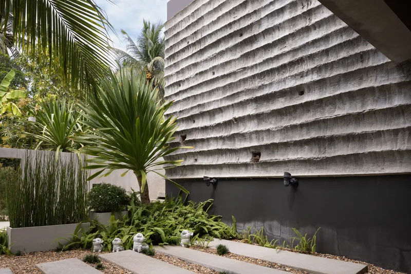 Ribbed concrete wall beside planted beds with palms and gravel under afternoon light