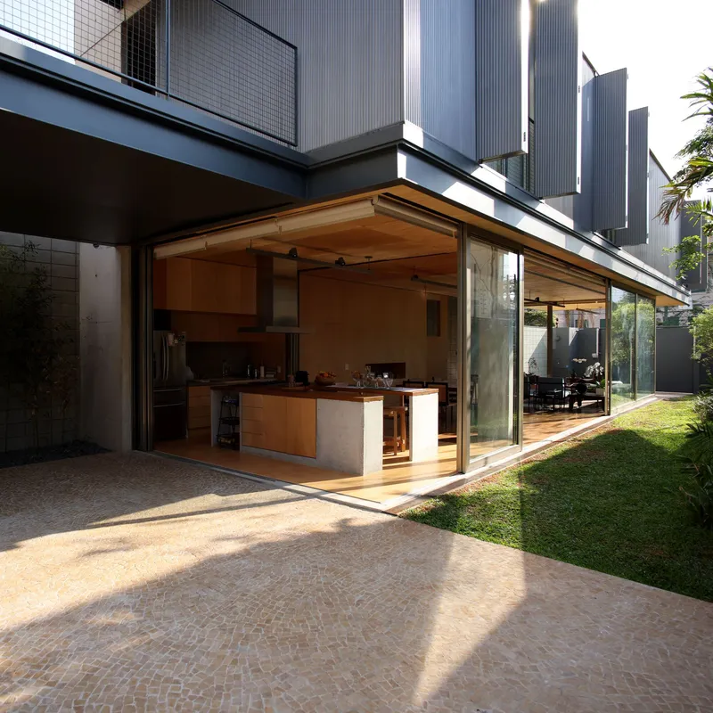 Open kitchen beneath timber-lined canopy extending from glass walls to brick-paved terrace at dusk