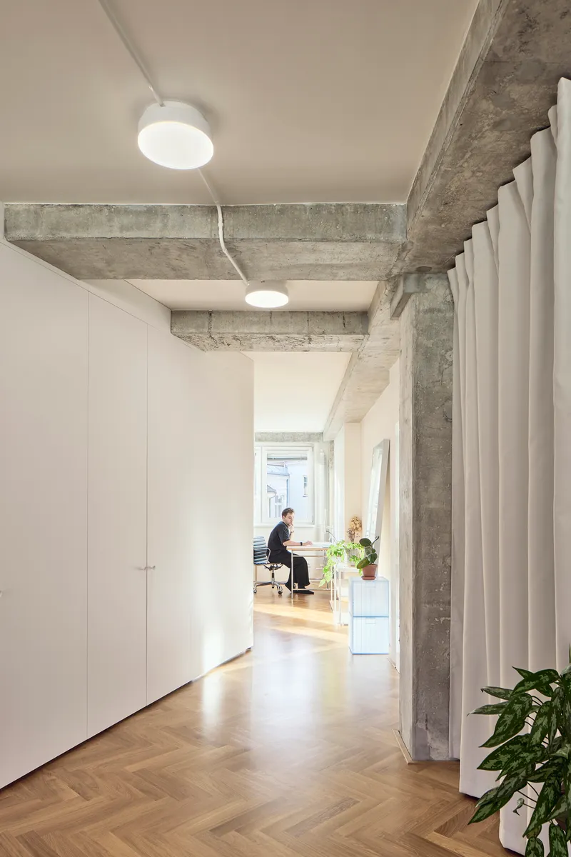 Hallway view showing exposed concrete beams and a figure working at a desk near the window