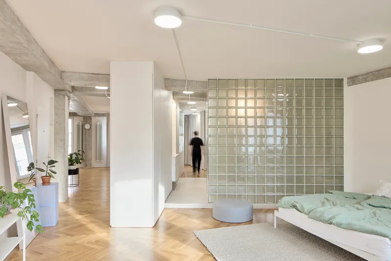 Bedroom with translucent glass block wall and a figure walking down the corridor beyond