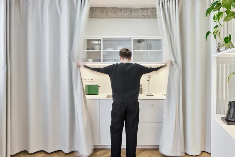 Person drawing back floor-to-ceiling curtains to reveal a compact white kitchen with concrete beam above