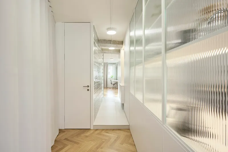 Corridor with fluted glass partition wall and herringbone wood floor transitioning to light tile