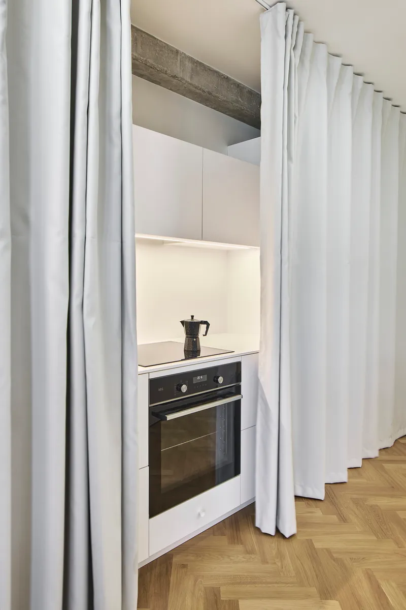 Built-in oven alcove concealed behind white fabric curtains and exposed concrete beam overhead
