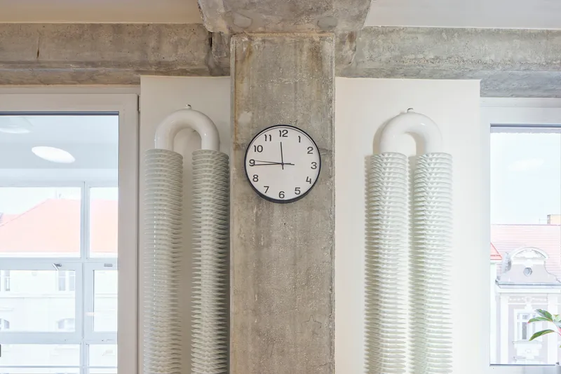 Exposed concrete column with ribbed air ducts flanking a clock in a white interior space