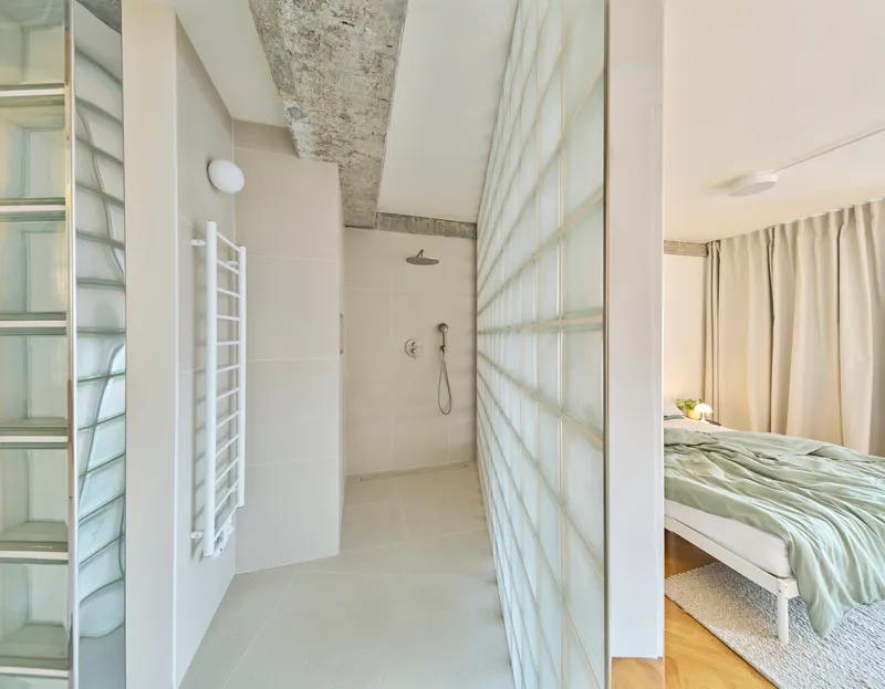 Bathroom with glass block wall and exposed concrete beam connecting to adjacent bedroom with curtain