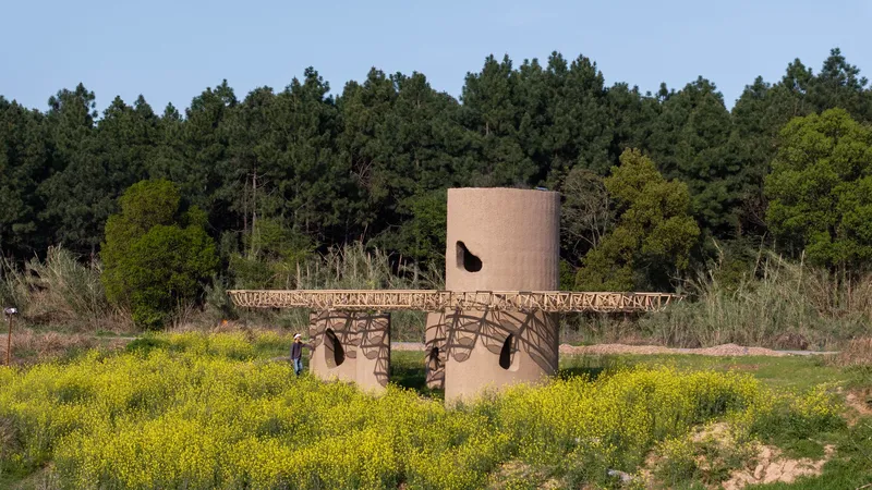 Cylindrical concrete tower with opening and timber canopy structure set among yellow wildflowers and dense forest backdrop