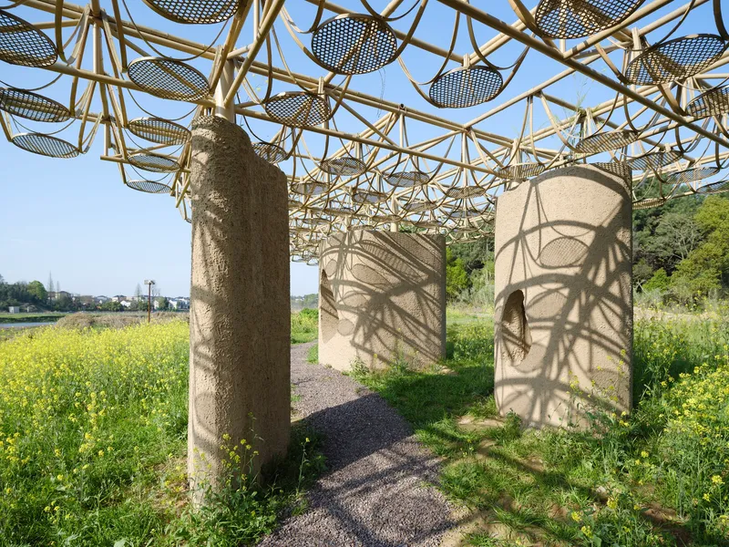 Bamboo lattice canopy supported by cylindrical columns casting dappled shadows on a gravel path through wildflowers