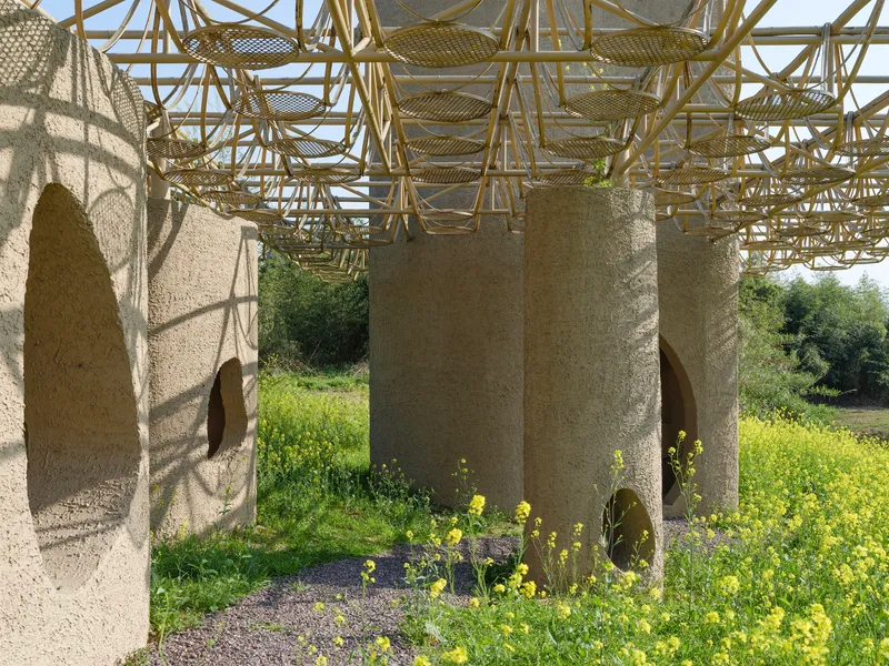 Cylindrical columns with teardrop-shaped openings beneath a bamboo lattice roof in a spring meadow