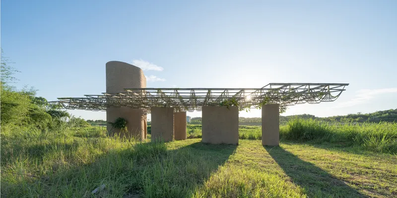 Rammed earth columns supporting a bamboo and wicker canopy structure in a grassy meadow at sunset